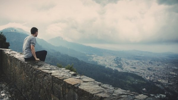 man sitting alone on concrete brick wall facing mountain and city under cloudy sky
